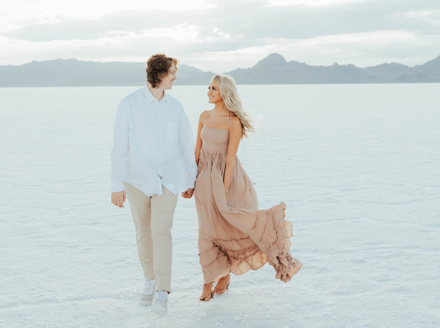 couple holding hands and walking toward the camera as bride to be's blush boho dress & hair are wind blown