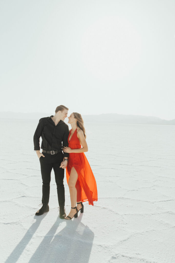 Couple standing close and smiling at each other during destination Salt Flats engagements, with the bride’s red dress flowing against the white landscape.