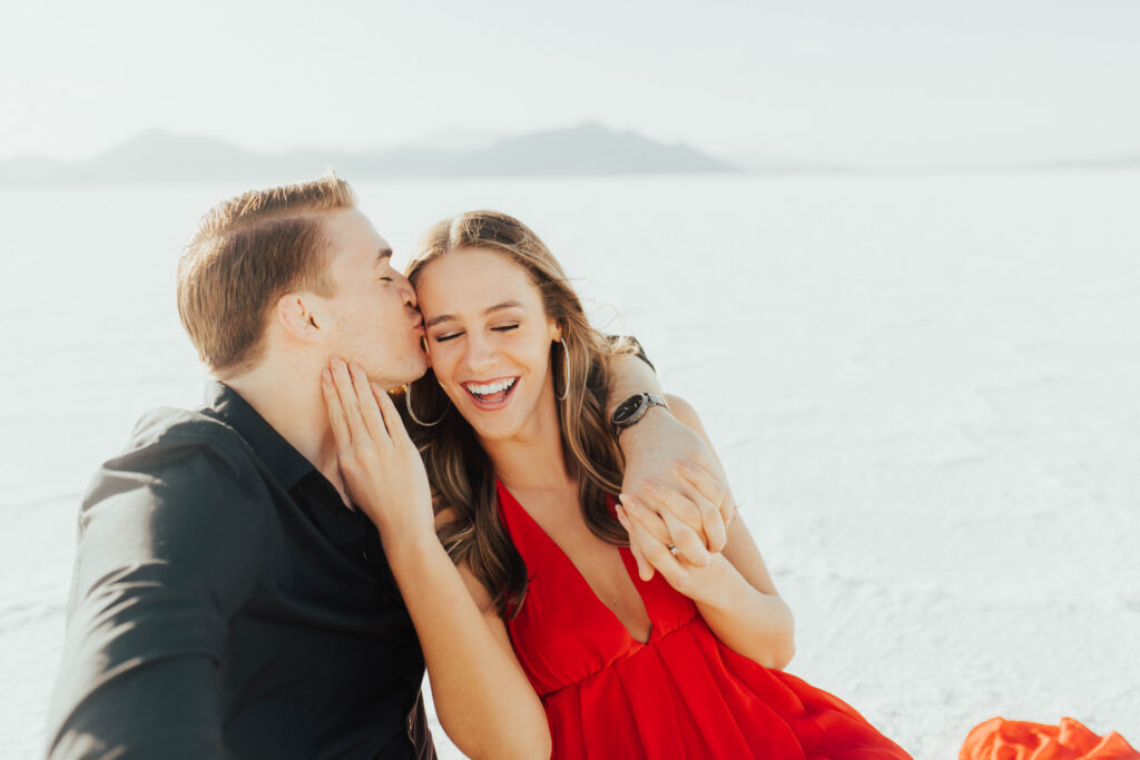 Groom kissing his fiancée’s cheek as she laughs in a red dress during their joyful destination Salt Flats engagements.