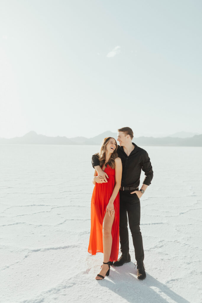 Groom holding bride from behind as she laughs in a bold red dress during their destination Salt Flats engagement session at Bonneville.