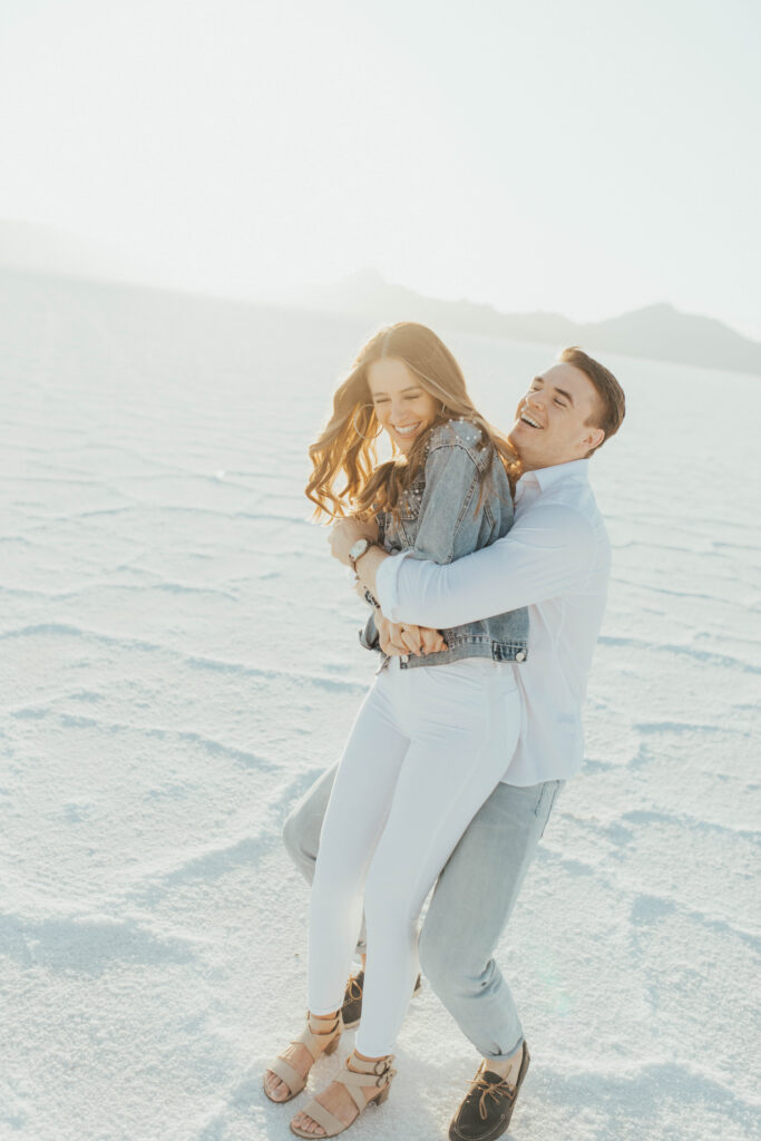 Groom wrapping bride in a playful hug during their destination Salt Flats engagements, sunlight glowing around them.