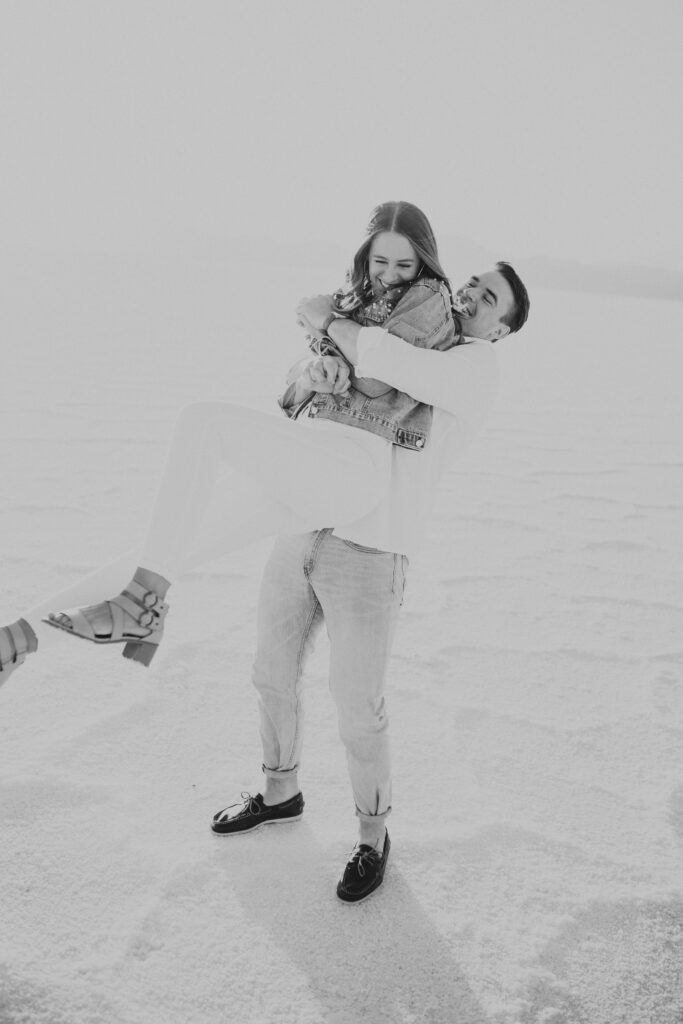 Black and white photo of groom lifting bride as they laugh during their destination Salt Flats engagements.