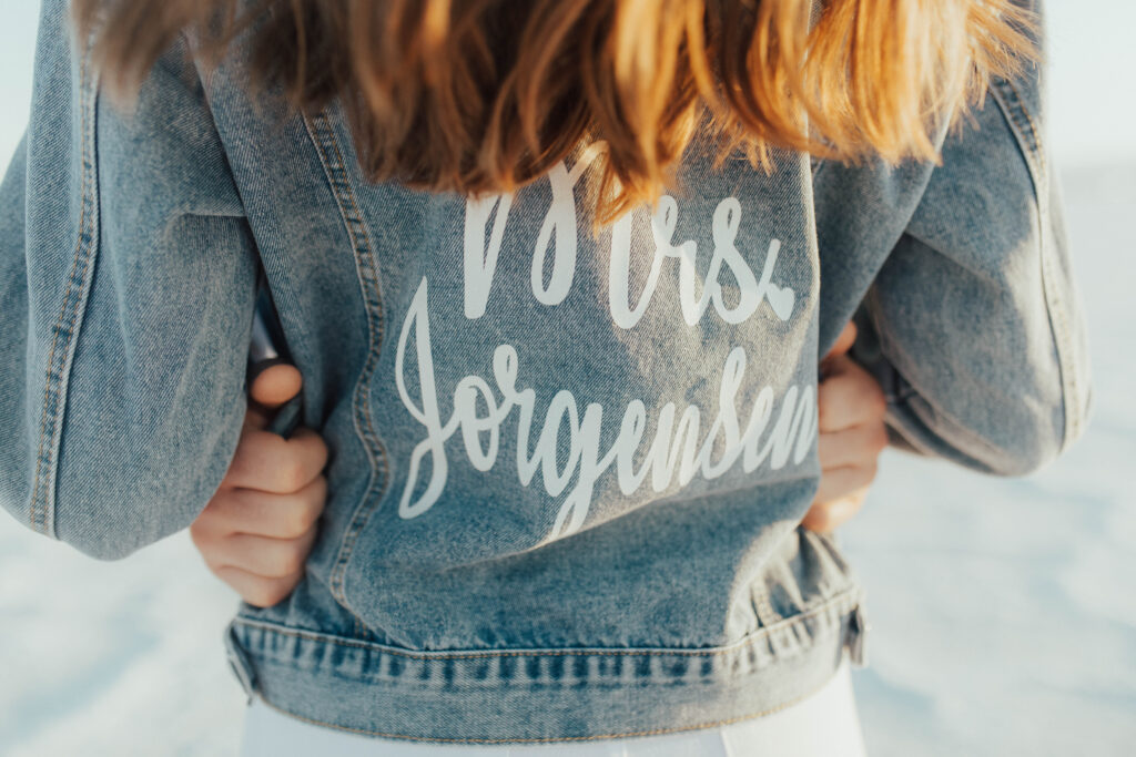 Close-up of custom “Mrs. Jorgensen” denim jacket during destination Salt Flats engagements, with groom’s hands wrapped around the bride.