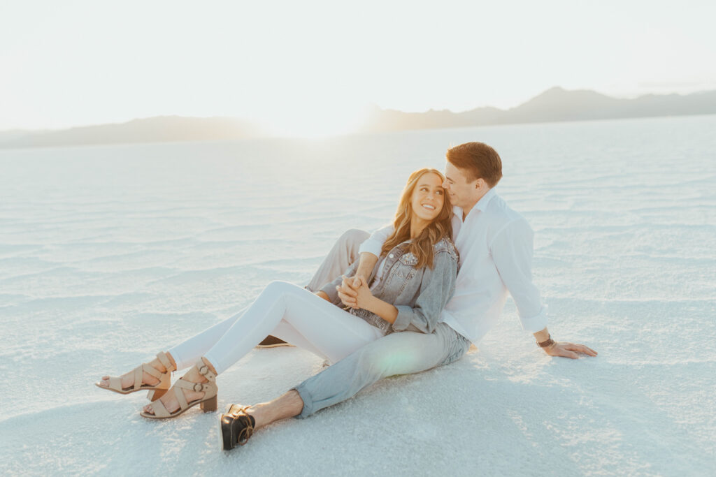 Bride smiling at her fiancé while sitting together during their destination Salt Flats engagements at sunset.